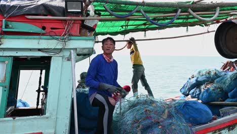 A-fisherman-with-a-cigarette-in-his-mouth-talking-and-gesturing-while-mending-a-net,-Pattaya-Fishing-Dock,-Chonburi,-Thailand