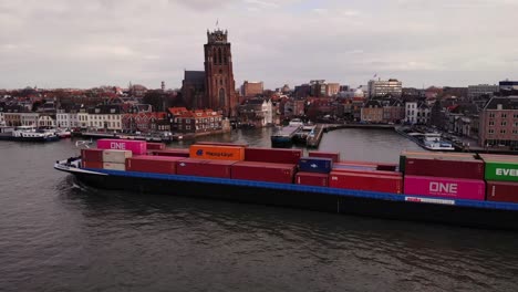 Aerial-Port-Side-View-Of-Sensation-Cargo-Ship-Going-Past-On-Oude-Maas-With-Our-Lady-of-Dordrecht-In-Background