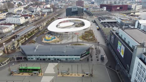 Bezirksgericht-Sandnes-Im-Hintergrund---Busbahnhof-Mit-Grünem-Bus-Und-Glasdachgebäude-Im-Vordergrund---Modern-Gestalteter-Stadtplatz-Mit-Spielplatz-Und-Eislaufarena-In-Der-Mitte---Norwegen-Aus-Der-Luft