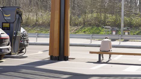 Hombre-Esperando-En-El-Banco-De-La-Estación-De-La-Autopista-Mientras-Se-Carga-Su-Coche-Eléctrico