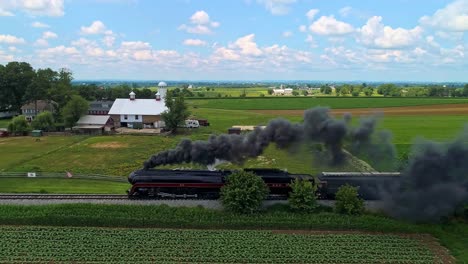 Una-Vista-Aérea-Paralela-De-Un-Antiguo-Tren-De-Pasajeros-De-Vapor-Que-Viaja-Con-Humo-Negro-A-Través-De-Tierras-De-Cultivo-En-Un-Día-Soleado