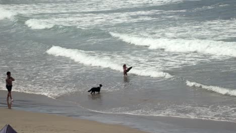 Niño-Pequeño-En-El-Océano-Con-Un-Tablero-De-Cuerpo-Y-Su-Perro-Negro-De-Pie-En-La-Playa