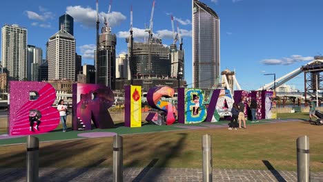 Sonniger-Tag-Am-Flussufer,-Touristen-Am-Wahrzeichen,-Buntes-Blockschild-Der-Stadt-Brisbane-Mit-Im-Bau-Befindlicher-Cross-River-Bridge-Im-Hintergrund,-Queensland,-Der-Sunshine-State,-Australien