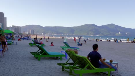 Video-Cinematográfico-De-La-Playa-Más-Hermosa-Y-Concurrida-De-Vietnam,-Da-Nang-Con-Gente-Sentada-En-Una-Silla-De-Playa,-Paseando-Por-La-Arena-Y-Divirtiéndose-En-El-Agua-Y-En-Una-Colina-Cercana