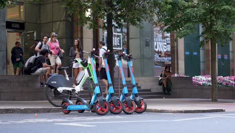 London-Canary-Wharf,-August-2022-Reihe-Von-Elektrorollern-Und-Fahrrädern-Parken-Am-Straßenrand-Und-Warten-Auf-Passagiere,-Während-Menschen-Vorbeigehen