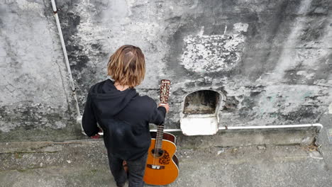 Foto-Superior-De-Un-Joven-Con-Una-Sudadera-Con-Capucha-Negra-Caminando-Hacia-Una-Guitarra-Clásica,-Luego-Tomándola-Con-Una-Mano-Y-Manteniéndola-Quieta,-En-Una-Estructura-Abandonada,-En-Un-Día-Nublado.