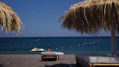 Mujer-En-Rojo-Camina-Por-El-Océano-En-La-Playa-De-Perissa-En-Santorini-Con-Arena-De-Lava-En-Grecia,-Europa