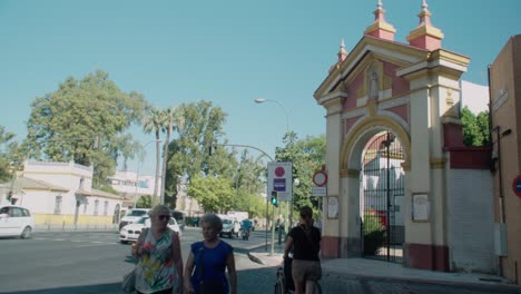 People-walk-past-arch-gate-to-convent-in-Seville,-Spain,-on-summer-day