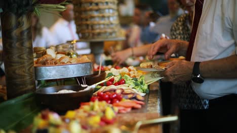 Smooth-shot-of-a-line-of-people-at-a-crowded-event-going-through-a-food-line-and-grabbing-a-variety-of-food-including-meats,-cheeses,-bread,-fruits,-and-vegetables