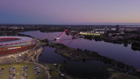 Toma-Aérea-Cinematográfica-De-Un-Estadio-Junto-Al-Río-Swan-Con-Movimiento-Hacia-Atrás-Y-Alrededor-Que-Revela-El-Horizonte-De-Perth-Al-Atardecer-Y-Un-Puente-Con-Tráfico-Moderado