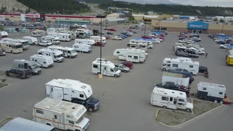 Imágenes-Aéreas-De-Un-Estacionamiento-De-Walmart-En-Whitehorse,-Canadá,-Que-Muestran-Muchos-Vehículos-Recreativos-Estacionados-Con-Autos-En-Movimiento,-Gente-Caminando,-Pájaros-Volando-Y-Montañas-En-El-Fondo