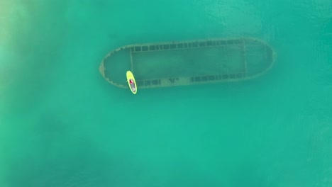 Birds-eye-view-of-woman-stand-up-paddle-over-rusty-shipwreck-in-clear-turquoise-water-by-tropical-island