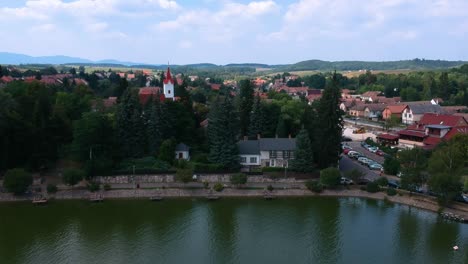 Flying-over-the-lakeshore-towards-the-town-center-of-Bánk,-with-the-mountains-of-Börzsöny-in-the-background-at-Nógrád-County,-Hungary