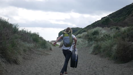 Cámara-Lenta-De-Un-Joven-Rubio-Corriendo-Alegremente-Hacia-El-Océano,-Con-Una-Mochila-Y-Una-Tapa-De-Guitarra,-En-Un-Camino-De-Arena-Rodeado-De-Hierba-Alta-En-La-Playa-De-Bethells,-En-Un-Día-Nublado-Y-Ventoso