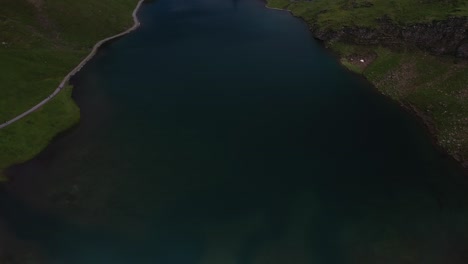 Drone-shot-panning-up-from-beautiful-and-colorful-green-fields-and-turquoise-blue-lake,-to-an-amazing-view-of-the-snow-covered-mountain-tops-of-Switzerland-during-a-sunny-summer-day