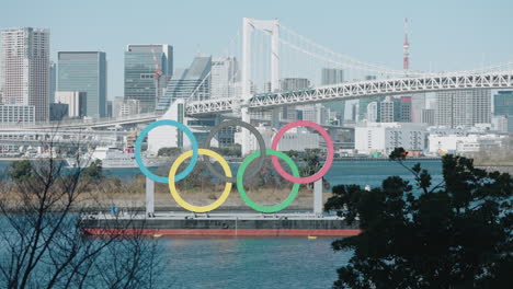 Denkmal-Der-Olympischen-Ringe-Im-Odaiba-Marine-Park-Mit-Segelboot,-Stadtbild,-Regenbogenbrücke-Und-Tokyo-Tower-Im-Hintergrund-In-Minato,-Tokio,-Japan