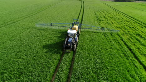 Drohnenaufnahme-Eines-Blauen-Traktors-Mit-Gelbem-Anhänger,-Der-Pflanzen-Auf-Der-Grünen-Wiese-Mit-Insektiziden-Besprüht