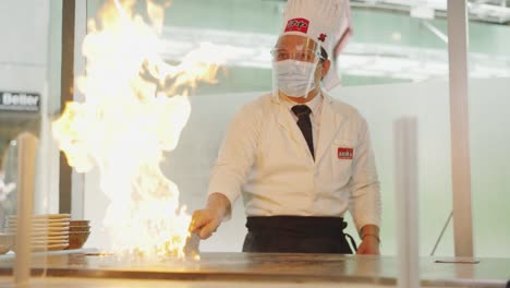 Teppanyaki-Show-Vor-Kunden-–-Erfahrener-Koch-Mit-Gesichtsmaske-Und-Gesichtsschutz-Mit-Schaber-Setzt-In-Einem-Asiatischen-Restaurant-Öl-In-Einer-Heißen-Bratpfanne-In-Brand