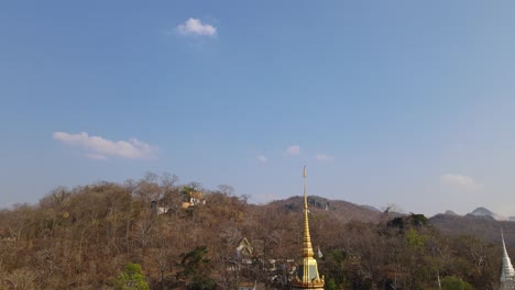 Wat-Phra-Phutthabat,-Saraburi,-Thailand,-ascending-aerial-shot-of-the-golden-Buddhist-temple,-sliding-to-the-left-revealing-brown-trees-in-summer,-a-hill-with-a-stairway-going-up-to-another-temple