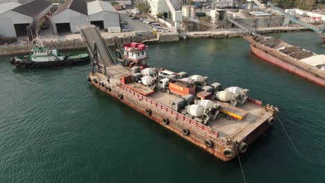 Barge-loaded-with-Concrete-mixer-trucks-pulled-to-port-by-a-Tugboat-in-Hong-Kong-bay,-Aerial-view