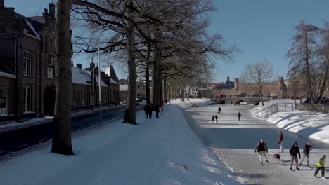 Paisaje-Nevado-Aéreo-De-Río-Congelado-Con-Portal-De-Barco-Medieval-Berkelpoort-En-El-Fondo-Y-Gente-Patinando-Sobre-Hielo-Y-Disfrutando-Del-Canal-Sólido-Contra-Un-Cielo-Azul