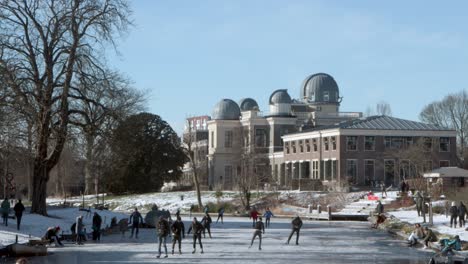 Gente-Patinando-Sobre-Hielo-En-Un-Canal-Congelado-Frente-Al-Observatorio-De-Leiden-En-Los-Países-Bajos-En-Un-Soleado-Día-De-Invierno-Durante-La-Pandemia