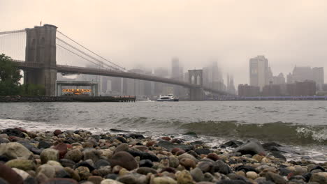 Wellen-Am-Ufer-Des-East-River-Mit-Brooklyn-Bridge-Und-Downtown-Manhattan-In-Der-Ferne,-New-York,-USA