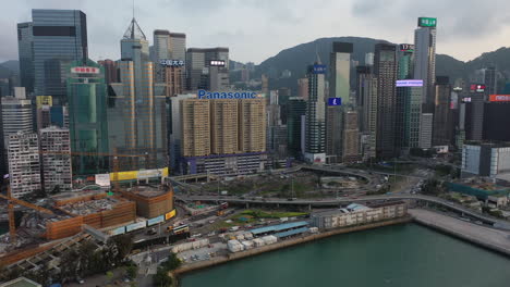 Vista-Aérea-De-Los-Edificios-Corporativos-Frente-Al-Mar-Central-De-Hong-Kong,-Tráfico-Nocturno-Y-Sitio-De-Construcción-En-El-Paseo-Marítimo,-Revelando-Disparos-De-Drones