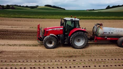 Vuelo-De-Drones-Cerca-Del-Tractor-Con-Remolque-Conduce-Sobre-El-Campo-Y-Vierte-Plantas-De-Lechuga-Jóvenes-Con-Agua