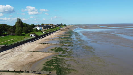 Un-Vuelo-De-Drones-Sobre-Una-Playa-Con-Viejos-Espigones-De-Madera-Y-Marismas,-Con-Casas-Detrás-De-Un-Dique-Marino-A-La-Izquierda,-En-El-Estuario-De-Las-Mareas-Con-Marea-Baja-En-Un-Día-De-Verano-Claro-Y-Caluroso