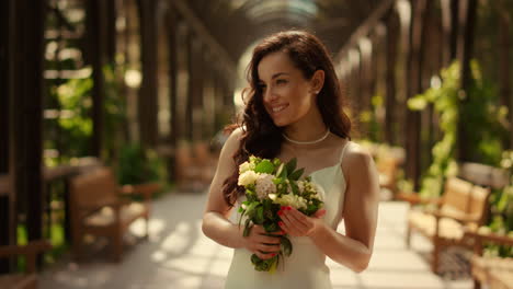 Charming-bride-posing-with-bouquet-under-arch.-Woman-feeling-happy-in-park