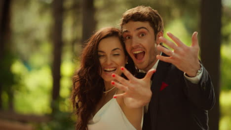 Positive-bride-and-groom-posing-on-wedding.-Couple-showing-rings-in-park