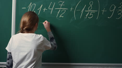 Back-view-of-school-girl-doing-math-task-on-blackboard-in-class.Student-writing