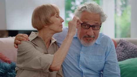Senior-woman-taking-off-eyeglasses-from-man.-Mature-couple-sitting-on-couch