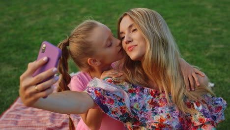 Girl-kissing-woman-in-garden.-Family-taking-selfie-on-mobile-phone-outdoor.