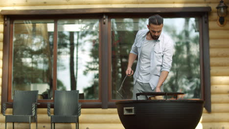 Focused-guy-using-poker-outdoors.-Man-chef-cooking-food-on-bbq-grill-near-house.