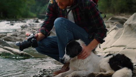 Photographer-showing-dog-photos-on-camera.-Hiker-sitting-at-river-with-dog