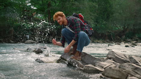 Male-traveller-taking-break-at-river-during-hike.-Redhead-man-splashing-water