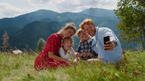 Positive-family-taking-selfie-on-cellphone-sitting-green-grass-mountain-hill.