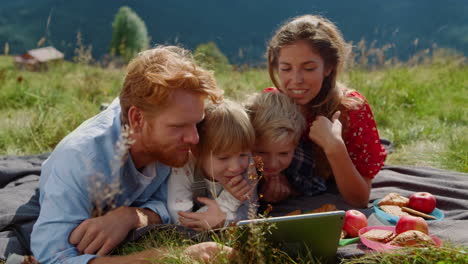 Family-looking-tablet-screen-lying-green-meadow-close-up.-Parents-kids-relaxing.