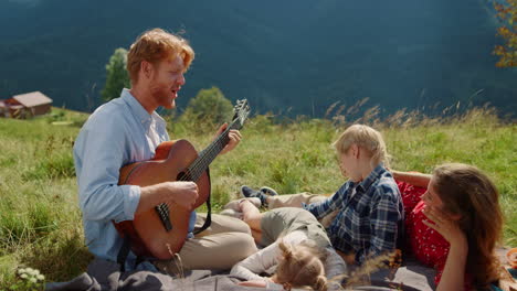 Father-playing-guitar-nature.-Happy-family-singing-sitting-blanket-at-picnic.