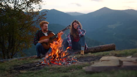 Young-couple-relax-camp-in-mountains.-Hiking-hipsters-play-guitar-at-campfire.