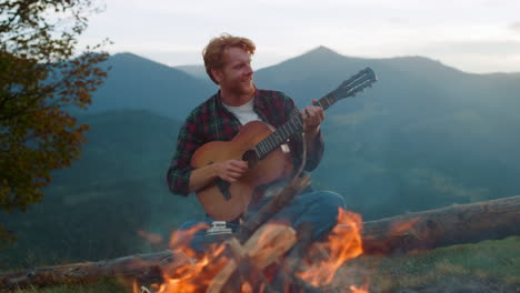 Happy-guy-sing-camp-song-in-mountains.-Positive-hipster-play-guitar-on-nature.