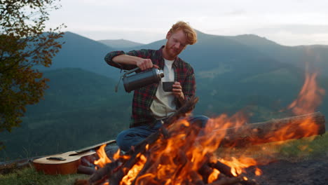 Relaxed-tourist-enjoy-camping-lifestyle-by-bonfire.-Chill-guy-drink-tea-outside.