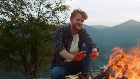 Smiling-traveler-warm-fire-on-camp.-Joyful-millennial-rest-in-forest-closeup.