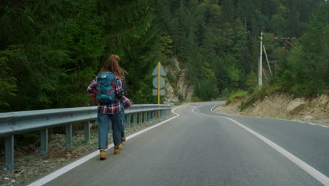 Couple-hitchhikers-walk-mountains.-Two-travelers-trekking-on-roadside-highway.