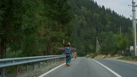Felices-Excursionistas-Caminando-Por-La-Carretera-En-El-Paisaje-De-Las-Montañas.-Dos-Amigos-Levantando-La-Mano