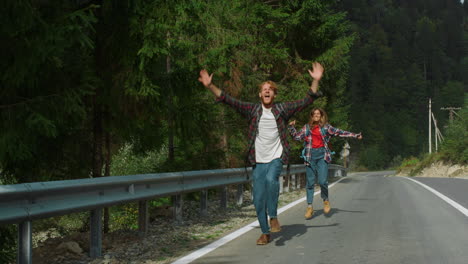 Overjoyed-hitchhikers-waving-hands-on-mountain-road.-Couple-jump-on-highway.