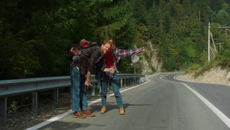 Two-travelers-waving-hands-catch-car-in-mountains.-Couple-standing-on-roadside.