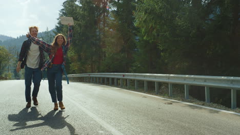 Couple-walk-forest-highway-in-mountains.-Hitchhikers-wait-car-wave-hands-outside
